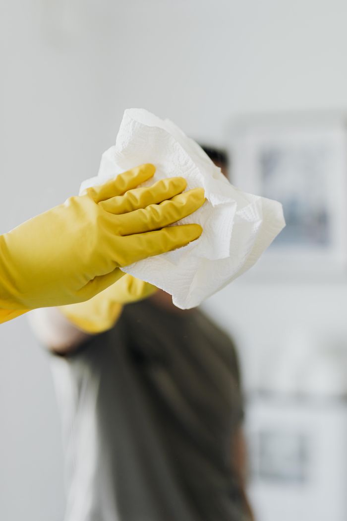 Homme anonyme portant des gants en latex jaunes et un t-shirt sombre, essuyant le miroir de la salle de bain avec une serviette en papier